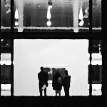 3 Menschen schauen sich in einer Galerie eine Bild an. Links und rechts stehen Pfeiler und Scheinwerfer beleuchten den Raum.